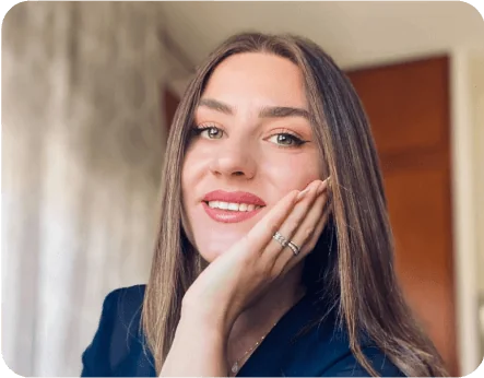 Young woman with long straight brown hair smiles, resting her hand against her cheek (showing two rings); soft indoor light, blurred curtains and a...