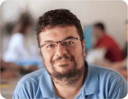 Bearded man with glasses smiles gently at the camera while seated, wearing a light blue polo; blurred indoor setting with people and desks visible in...