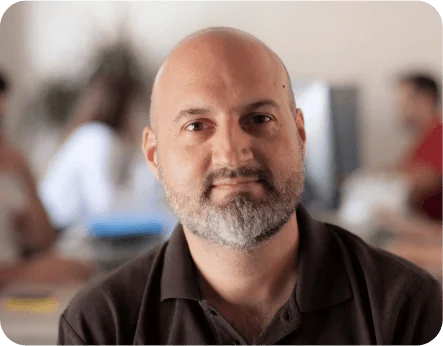 Bald man with trimmed salt-and-pepper beard looks directly at camera with a slight smile, wearing a dark polo, seated in a softly lit office with...