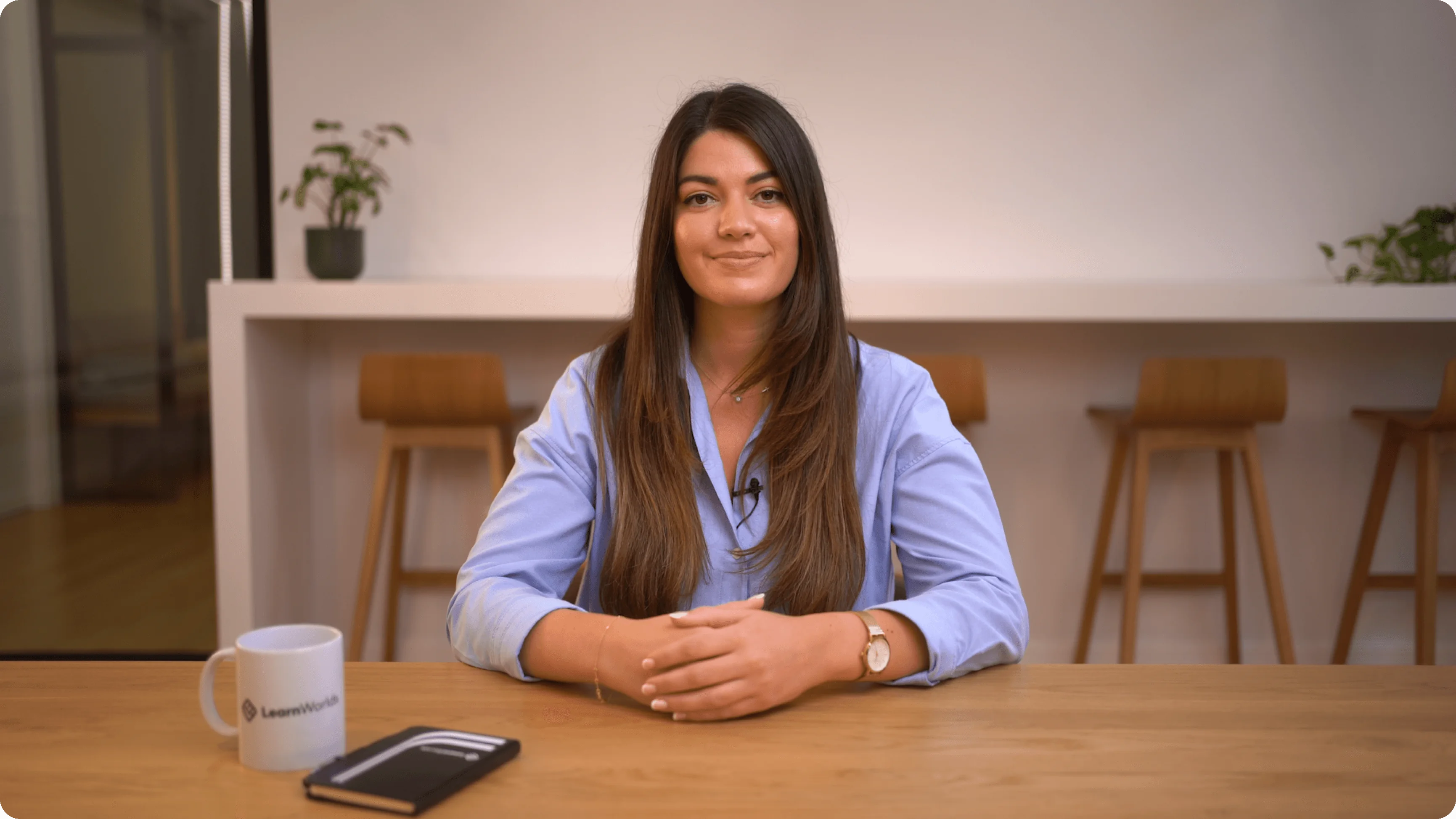 Young woman sits smiling with hands folded on a wooden table; wearing a blue shirt and lapel mic; modern workspace with bar stools, plants, a mug...