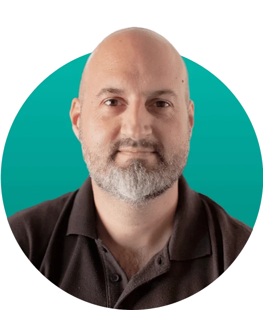 Bald man with trimmed gray beard gazes directly at the camera with a neutral expression against a teal circular backdrop, wearing a dark polo shirt.