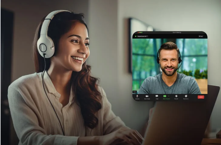 Woman with headphones smiles and types on a laptop while a man appears in a Zoom window, wearing a headset against a bright indoor background.