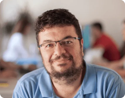 Bearded man wearing glasses and a light-blue polo smiles gently at the camera, seated in a softly lit room with blurred people and tables in the...