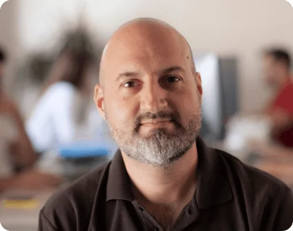 Bald, bearded man gazes into the camera with a slight smile, seated in a softly lit open office; blurred coworkers and desks form a neutral...
