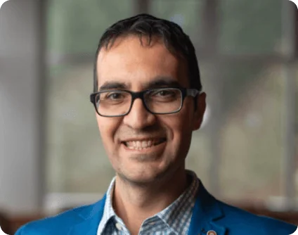 Man wearing black glasses smiles at the camera, dressed in a blue blazer and checkered shirt, standing indoors in front of a softly blurred window...