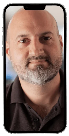 A bald man with a salt-and-pepper beard looks at the camera with a neutral expression; wearing a dark polo, he is framed in a smartphone screen...