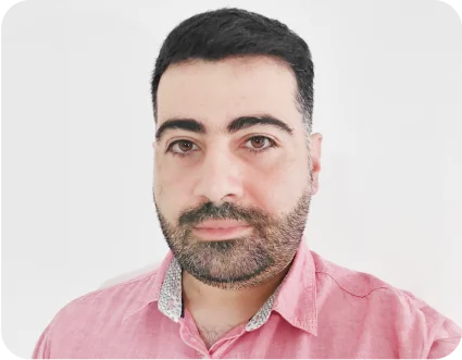 Adult man in a pink collared shirt gazes directly at the camera; light beard, neutral expression against a plain white, softly lit background.
