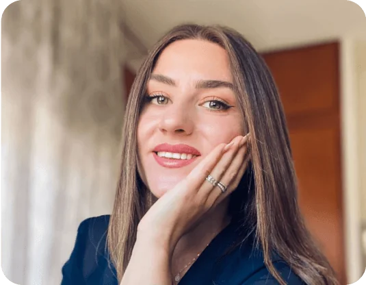 Woman smiling, resting her chin on her hand; wearing rings and a navy top; long straight hair; softly lit indoor room with blurred curtains and a...