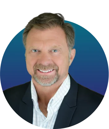 A middle-aged man with short graying hair and trimmed goatee smiles at the camera, wearing a dark blazer and white collared shirt against a circular...