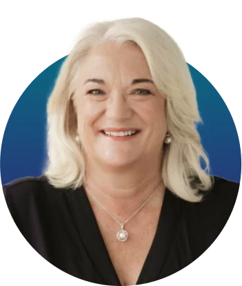 A smiling middle-aged woman with white hair, wearing a black blouse, pearl stud earrings and a silver pendant necklace, posed in a circular...