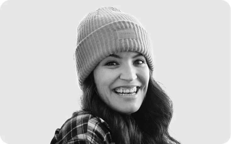 Woman smiling broadly, looking over her shoulder; wearing a ribbed beanie with a small rectangular label (text illegible), plaid shirt, long hair,...