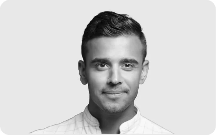 Young man looking directly at the camera with a slight smile, short coiffed hair, wearing a light patterned collared shirt against a plain pale-gray...