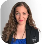 Person with long curly brown hair smiles at camera, wearing a black blazer over a blue top with a small white wing-like emblem; plain light-gray...