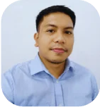 A man faces the camera with a neutral expression, wearing a light-blue button-up shirt; posed head-and-shoulders against a plain pale wall in soft,...