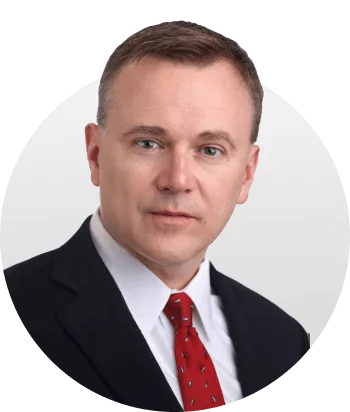 A middle-aged man faces the camera with a neutral expression, wearing a dark suit, white shirt and red tie; head-and-shoulders portrait on a light...