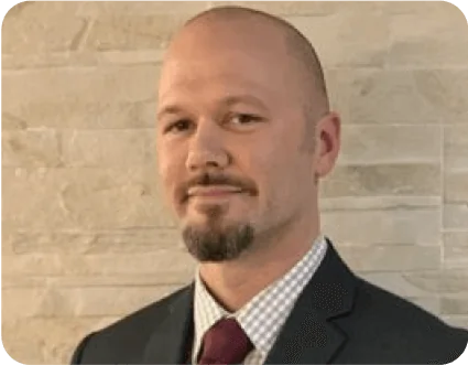 Adult man with a shaved head and goatee, wearing a dark suit, checked shirt and maroon tie, posing with a slight smile while facing the camera...