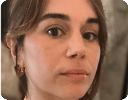 Close-up of a woman’s face looking straight at the camera; neutral expression, brown hair tucked behind one ear with a small hoop earring; blurred...
