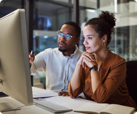 A desktop monitor displays content as two colleagues review it; a man points at the screen while a woman watches attentively, seated at a desk with...