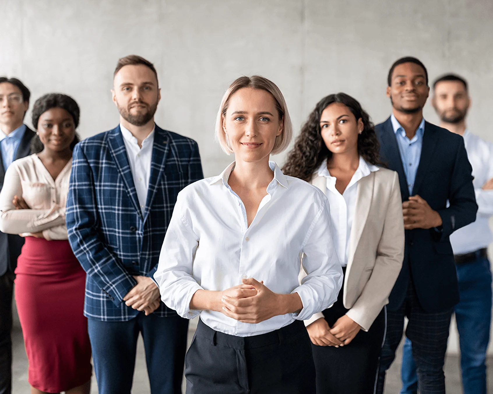 A woman stands confidently at front, hands clasped; six diverse colleagues stand behind her in business attire, forming a professional team against a...